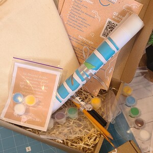 May include: A craft kit in a cardboard box containing a rolled canvas with a blue and white pattern, a small plastic bag with three paint pots, a set of small paint containers, two pencils, a black marker, and printed instruction sheets.