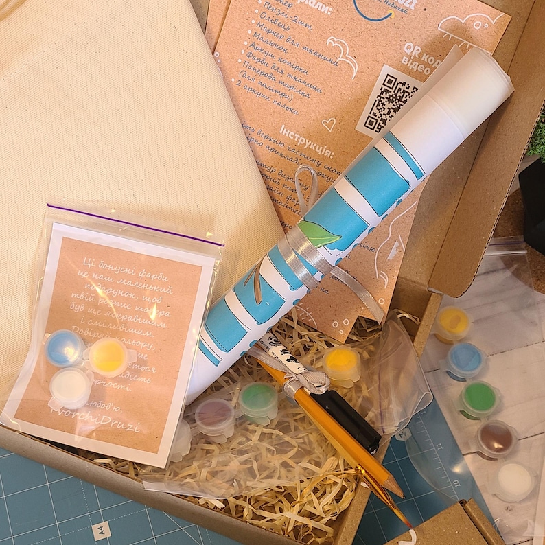 May include: A craft kit in a cardboard box containing a rolled canvas with a blue and white pattern, a small plastic bag with three paint pots, a set of small paint containers, two pencils, a black marker, and printed instruction sheets.