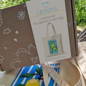 May include: A brown cardboard box labeled Lemons Creative kit for painting a tote bag with a graphic of a lemon-printed tote bag. Below the box sits a cream-colored canvas tote bag featuring a painted design of yellow lemons on blue stripes.