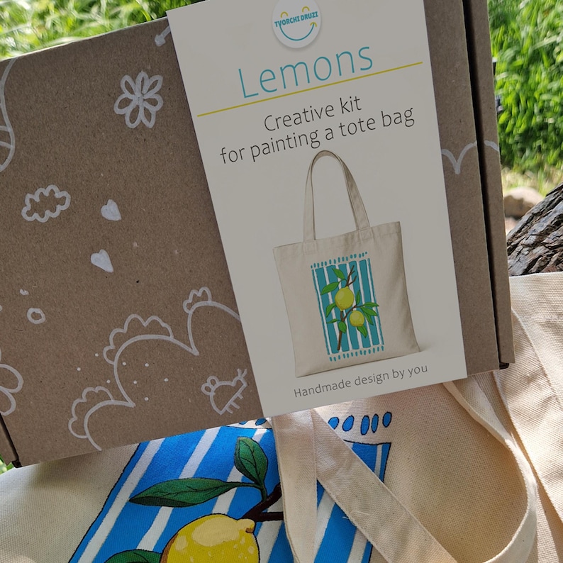 May include: A brown cardboard box labeled Lemons Creative kit for painting a tote bag with a graphic of a lemon-printed tote bag. Below the box sits a cream-colored canvas tote bag featuring a painted design of yellow lemons on blue stripes.