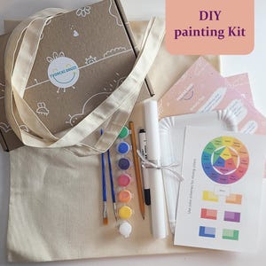May include: A DIY painting kit featuring a canvas tote bag, paintbrushes, paint pots, and a colour mixing guide. The kit includes a brown cardboard box with a logo, a white canvas roll, and a pencil. The text "DIY painting Kit" is visible.