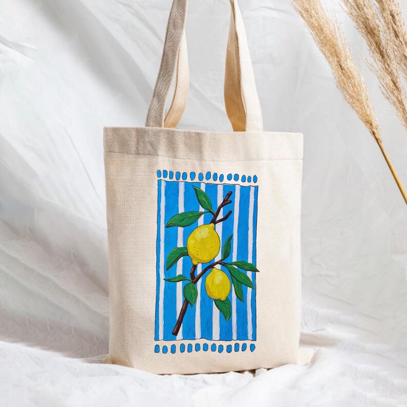 May include: A beige canvas tote bag featuring a rectangular graphic print. The design displays a branch with two yellow lemons and green leaves centered against a background of vertical blue and white stripes, framed by blue dots.