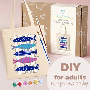 Puede incluir: Una bolsa de tela beige con un diseño estampado de cinco peces estilizados en azul, turquesa y rosa, acompañada de una caja de cartón etiquetada Sardines Creative kit for painting a tote bag, dos pinceles y seis pequeños botes de pintura.