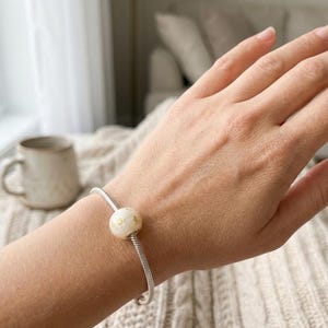 Puede incluir: Una pulsera de cadena de serpiente en tono plateado que presenta una cuenta redonda de color blanco roto con pequeñas motas doradas. La pulsera está asegurada alrededor de la muñeca, mostrando la textura metálica de la cadena y el acabado liso.