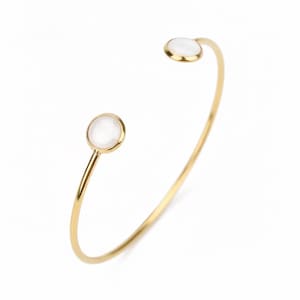 Op de afbeelding: Een goudkleurige open armband met een dunne, gepolijste metalen band. Aan elk uiteinde van de armband bevindt zich een ronde zetting met een gladde, ondoorzichtige witte steen of inleg, wat zorgt voor een gebalanceerd en symmetrisch ontwerp.