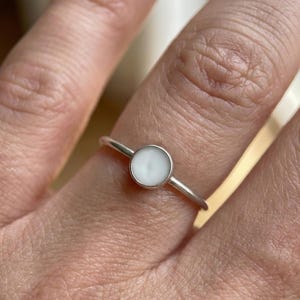 Op de afbeelding: Een zilveren ring met een ronde, witte, ondoorzichtige steen in een zetting. De ring heeft een slanke band en een gepolijste afwerking. De steen is ongeveer 0,6 cm in diameter, wat een minimalistische uitstraling geeft.