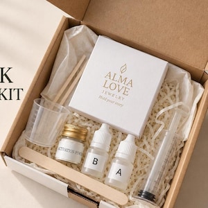 Könnte beinhalten: Ein brauner Karton mit einer weißen Schmuckschachtel mit der Aufschrift ALMA LOVE JEWELRY Hold your story, einem kleinen Glas Aktivierungspulver, zwei Tropfflaschen mit der Aufschrift A und B, einem Messbecher, einer Spritze und einem Holzstab.