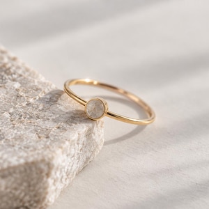 Pode incluir: Um anel fino de ouro com uma pequena pedra circular de cor clara engastada num bisel. A banda é lisa e polida, apresentando um design simples que destaca o contraste entre o acabamento metálico dourado e a pedra de tom pálido.