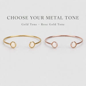 Op de afbeelding: Twee open armbanden, één in goudkleur en één in roségoudkleur, elk met een ronde witte steen aan beide uiteinden. De tekst CHOOSE YOUR METAL TONE, Gold Tone en Rose Gold Tone staat boven de armbanden.