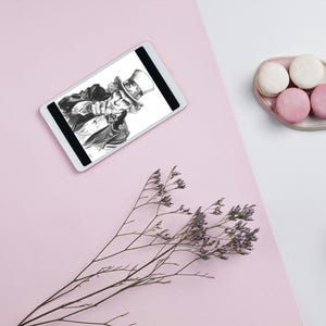 Könnte beinhalten: Ein digitales Tablet zeigt eine Schwarz-Weiß-Illustration einer Person mit Zylinder. Das Bild befindet sich auf einem rosa und weißen Hintergrund, mit Makronen und einer Tasse Kaffee. Getrocknete Blumen sind ebenfalls vorhanden.