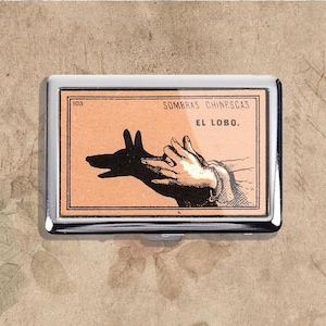 May include: A silver metal cigarette case with a rectangular orange insert. The insert features a black silhouette of a dog's head and hand shadows, with the text "SOMBRAS CHINESCAS EL LOBO."
