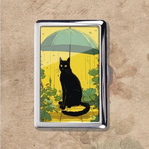 May include: A silver metal cigarette case featuring a black cat illustration under a green umbrella. The cat sits on a yellow background with green foliage. The case has a rectangular shape.