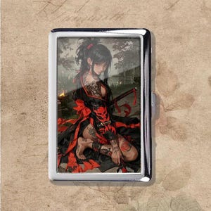 May include: A silver cigarette case featuring a detailed illustration of a woman in a black and red kimono, holding a katana. The artwork includes tattoos and a red skull mask, set against a blurred outdoor background.