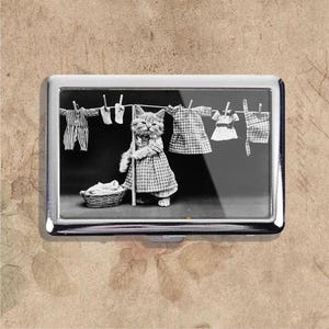 May include: A silver cigarette case with a monochrome image of a kitten in a checkered dress, holding a broom, with miniature clothes on a washing line. A basket of laundry sits at the kitten's feet, evoking a vintage aesthetic.