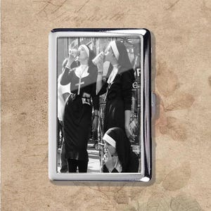 Puede incluir: Un pitillera plateada con una imagen en blanco y negro de monjas bebiendo y fumando. La pitillera tiene forma rectangular con esquinas redondeadas y un cierre lateral.
