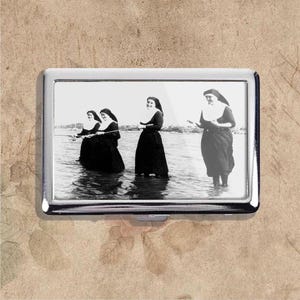May include: A silver cigarette case featuring a black and white image of four nuns standing in water. Three nuns are holding fishing rods, while the fourth nun is gesturing with her hands. The nuns are wearing traditional habits.
