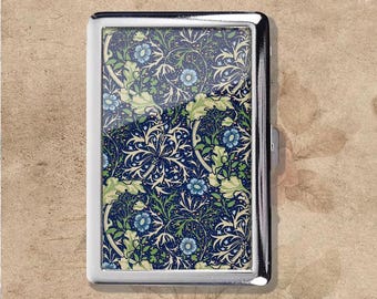 Puede incluir: Un estuche rectangular para cigarrillos con acabado plateado. Presenta un detallado diseño floral en azul, verde y blanco sobre un fondo azul oscuro. El diseño está enmarcado por un borde metálico pulido y tiene un cierre seguro.