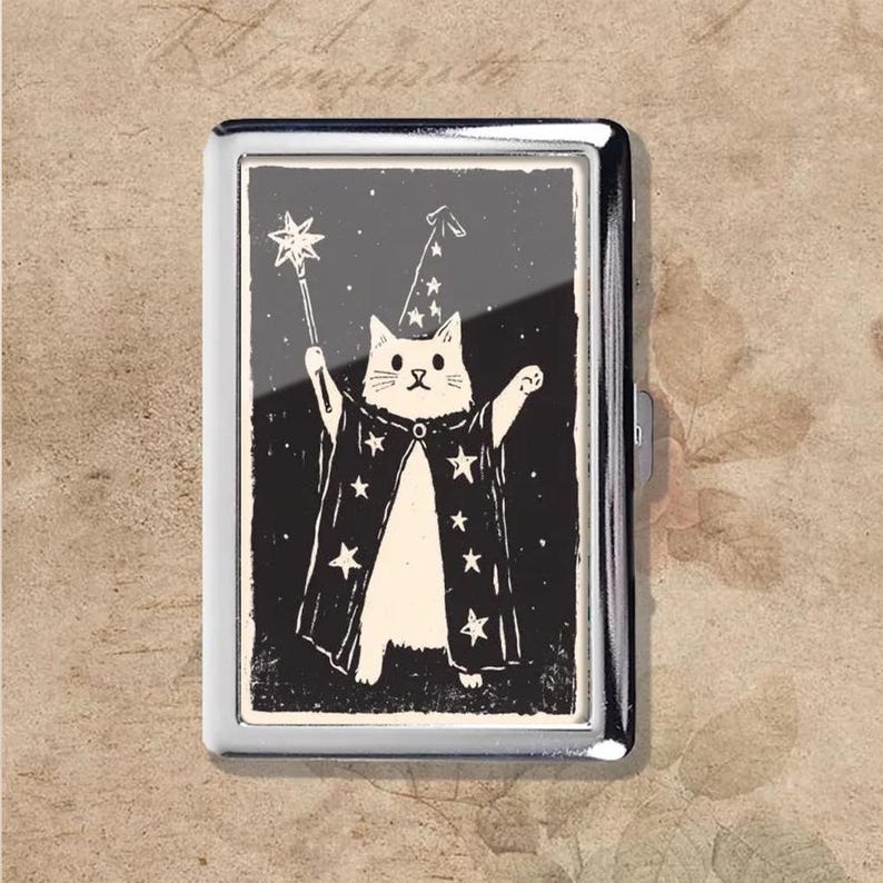 May include: A silver metal cigarette case featuring a black and white illustration of a cat dressed as a wizard. The cat wears a pointed hat and cape with stars, holding a wand with a star on the end.