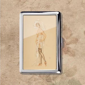 Puede incluir: Un estuche de cigarrillos de metal plateado con una ilustración vintage de una mujer con un vestido transparente y medias. La obra de arte está en tonos suaves y apagados, con un fondo color crema. El estuche tiene forma rectangular y cierre de bisagra.