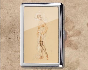 Puede incluir: Un estuche de cigarrillos de metal plateado con una ilustración vintage de una mujer con un vestido transparente y medias. La obra de arte está en tonos suaves y apagados, con un fondo color crema. El estuche tiene forma rectangular y cierre de bisagra.