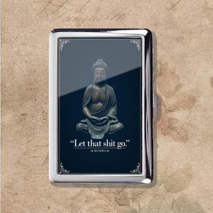 Puede incluir: Un pitillera plateada con un fondo azul oscuro que presenta una estatua de Buda sentado. La pitillera tiene detalles decorativos en las esquinas y la cita "Let that shit go." atribuida a Buda.