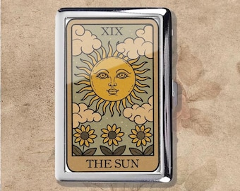 Puede incluir: Un estuche de cigarrillos plateado con un diseño de carta del tarot. La carta representa un sol sonriente con rayos, nubes y girasoles, con el texto "THE SUN" en la parte inferior. La carta está etiquetada como "XIX" en la parte superior.