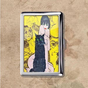 May include: A silver metal cigarette case featuring a colorful illustration. The artwork depicts a black cat with green eyes, a woman with red lipstick, and abstract faces on a yellow background.