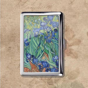 May include: A silver metal cigarette case featuring a painting of blue irises with green foliage. The artwork is set against a background of yellow and orange hues. The case has a rectangular shape and a clasp closure.