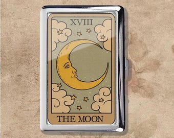 May include: A silver metal cigarette case featuring a tarot card design. The card depicts a crescent moon with a face, surrounded by clouds and stars, with the text "THE MOON" at the bottom.