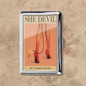 Puede incluir: Un estuche de cigarrillos de metal plateado con un gráfico retro. El gráfico presenta el texto "SHE DEVIL" sobre una ilustración de piernas con tacones y medias rojas, con una cola de diablo. La frase "She's coming for your soul" está debajo.