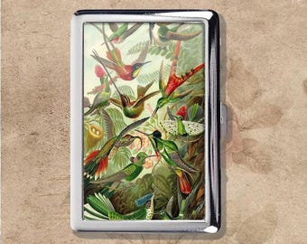 May include: A silver cigarette case with a detailed illustration of hummingbirds. The artwork shows various species of hummingbirds in flight, with vibrant green, red, and yellow colours set against a backdrop of foliage.