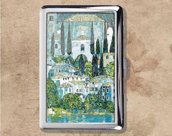 May include: A silver cigarette case with a painted landscape scene. The artwork shows a village with white buildings, tall trees, and a lake, under a blue sky. The case has a shiny, metallic finish.