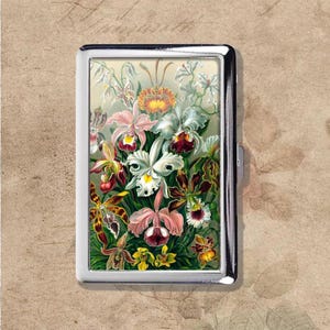 May include: A silver metal cigarette case featuring a colorful floral design. The artwork showcases a variety of orchids in shades of pink, white, yellow, and burgundy, set against a light green background. The case has a rectangular shape with rounded corners.