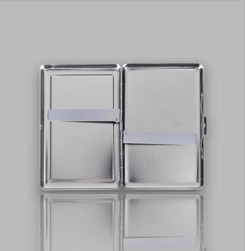 May include: Open, silver-colored cigarette case with a rectangular shape. The case is divided into two compartments, each with a silver band to hold items in place. The case is open, revealing the interior, and has a reflective surface.