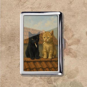 Puede incluir: Un estuche de cigarrillos plateado con una pintura de dos gatos en un tejado. Uno es negro, el otro es naranja, y ambos fuman cigarrillos. El fondo muestra un cielo azul y un tejado de tejas rojas.