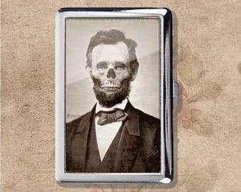 Pode incluir: Um porta-cigarros prateado com uma imagem em tons sépia de Abraham Lincoln com um crânio sobreposto. A imagem é emoldurada dentro do estojo retangular, que tem uma superfície brilhante e reflexiva. O estojo tem um fecho lateral.