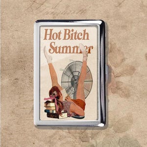 May include: A silver cigarette case with a retro illustration. The image depicts a woman with her legs raised, a fan, and the words "Hot Bitch Summer" in brown. The case has a shiny metal finish.