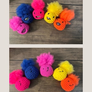 Op de afbeelding: Een verzameling kleurrijke, handgemaakte haakwezens. Elk wezen heeft een andere kleur, waaronder blauw, roze, geel en oranje, met een pluizige bovenkant en een lachend gezicht. De wezens zijn gerangschikt op een houten oppervlak.