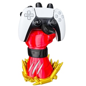 Può includere: Una scultura di mano rossa e muscolosa con un cinturino nero che tiene un controller per videogiochi bianco e nero. La base della mano rossa è circondata da diversi oggetti dorati a forma di proiettile e la mano presenta tre segni di artigli bianchi.