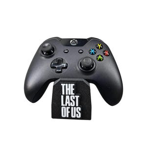 Könnte beinhalten: Ein schwarzer kabelloser Videospiel-Controller auf einem schwarzen Ständer. Der Ständer ist auf der Vorderseite mit dem weißen Text THE LAST OF US bedruckt. Der Controller verfügt über zwei Analogsticks, ein Steuerkreuz und vier farbige Aktionstasten.