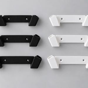 Pode incluir: Um conjunto de seis suportes de parede dispostos em duas colunas. Três suportes são preto fosco e três são branco fosco. Cada suporte apresenta uma barra horizontal com duas ranhuras anguladas abertas nas extremidades e dois orifícios.