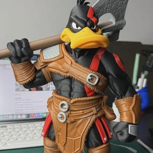 May include: A black duck figurine with a yellow beak, wearing brown leather-like armour with red straps and a red mark over one eye. The figure holds a large grey axe over its shoulder and wears brown boots, featuring detailed textures and moulded features.