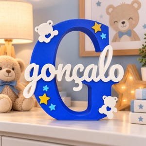 Könnte beinhalten: Ein großer, blauer, dreidimensionaler Buchstabe G mit dem weißen, kursiven Namen gonçalo in der Mitte. Der Buchstabe ist mit zwei weißen Teddybären-Silhouetten mit Herz-Ausschnitten, zwei gelben Sternen und zwei hellblauen Sternen verziert.