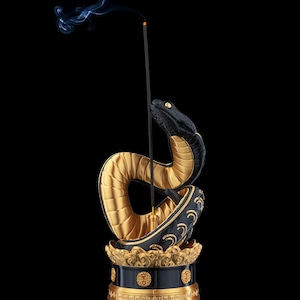 Pode incluir: Um porta-incenso preto e dourado com uma escultura de serpente enrolada sobre uma base circular com padrões decorativos. Uma vareta de incenso preta fina está na vertical no centro, emitindo um fio de fumo cinzento contra um fundo preto.