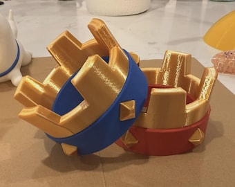 Può includere: Due corone stampate in 3D con una parte superiore in oro metallizzato e punte rettangolari alte. Una corona ha una base blu solida e l'altra una base rossa solida. Ogni base è decorata con piccoli borchie dorate a forma di piramide.