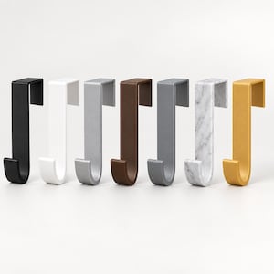 Op de afbeelding: Een set van zeven deurhaken op een rij. De haken hebben verschillende afwerkingen, waaronder matzwart, wit, zilver, bruin, donkergrijs, een wit marmerpatroon en een metallic gouden afwerking, elk met een standaard J-vormig ontwerp.