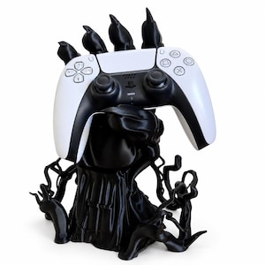 May include: A black, clawed hand stand holding a white and black video game controller. The base of the stand features intricate, root-like structures extending outwards, providing a stable foundation for the gaming accessory display.