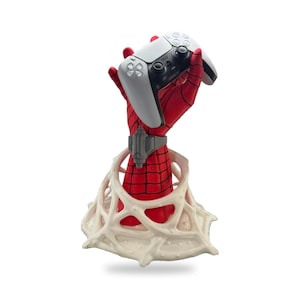 Puede incluir: Una escultura de mano roja y negra texturizada que emerge de una base blanca similar a una telaraña, con un accesorio de muñeca gris y sosteniendo un mando de videojuego blanco y negro. La mano presenta un patrón de rejilla en la superficie roja.