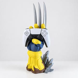 Può includere: Una scultura a forma di mano guantata gialla e blu con tre lunghi artigli argentati rivolti verso l'alto. La mano sostiene un controller per videogiochi bianco e nero. La base della scultura è formata da rocce grigie frastagliate per stabilità.