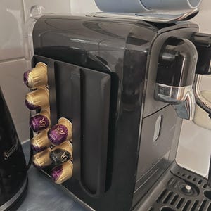 Pode incluir: Uma máquina de café preta com um suporte lateral para cápsulas contendo cápsulas de café douradas e roxas. A máquina possui um bico cromado, uma bandeja de gotejamento com luz indicadora vermelha e um acabamento brilhante.